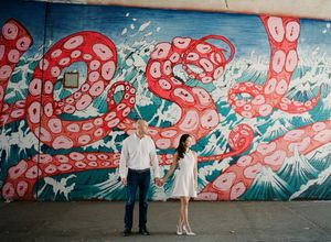 Couple in white holding hands in front of a vibrant red octopus street mural wall.