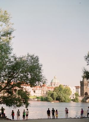 Couples and tourists stand along the Vltava River bank with Prague's historic architecture and domed buildings in the background.
