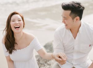 Happy Asian couple laughs joyfully while holding hands on a sunny beach during their engagement photo session.