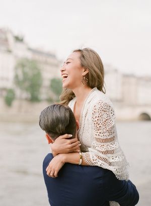 Joyful woman in crochet lace top laughs while being lifted by her partner along the Seine River in Paris.
