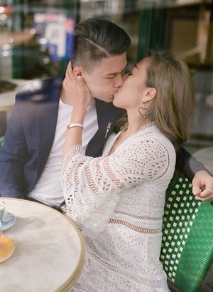 Romantic couple shares a kiss at a Parisian café, woman in white crochet dress with heart earrings.