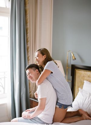 Laughing couple enjoys a playful moment together in a bright Parisian hotel bedroom with blue-gray curtains.