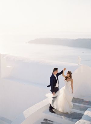 Bride and groom dancing on white Santorini rooftop steps with stunning caldera sea view.