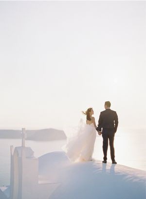 Bride and groom holding hands gazing at the breathtaking Santorini caldera from a white rooftop.