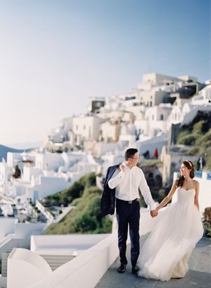 Couple walking hand in hand through iconic white-washed Santorini village steps at golden hour.