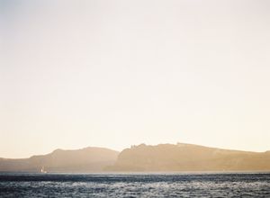Golden hour seascape with silhouetted coastal mountains and a lone sailboat on tranquil waters.
