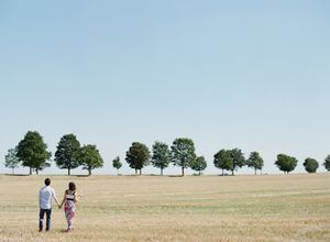 A couple holds hands walking across a vast golden field toward a row of green trees.