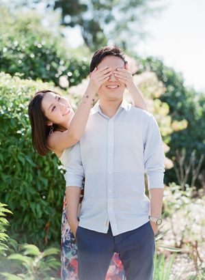 A woman playfully covers a smiling man's eyes from behind in a sunny garden setting.