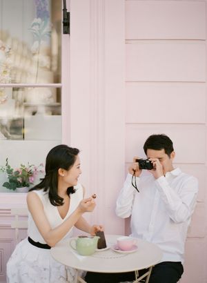 A couple enjoys a cafe date as a man photographs a laughing woman against a pink pastel wall.