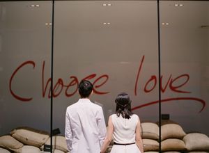 A couple stands before a glass window displaying the red graffiti phrase Choose Love.