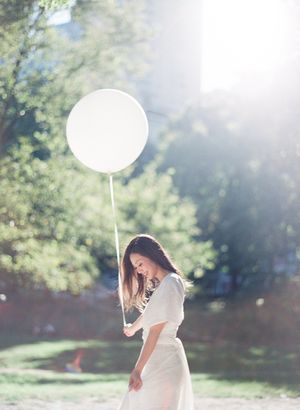 Woman in white dress holding a large white balloon in a sunlit park setting.