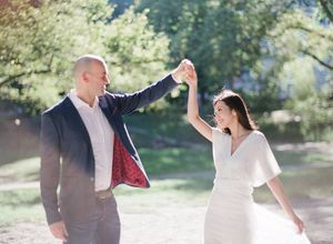 Joyful couple dancing in Central Park, man twirling woman in elegant white wrap dress.