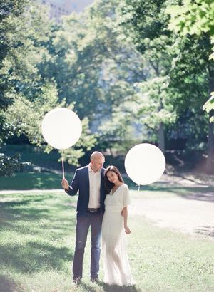 Engaged couple holding oversized white balloons while posing in a lush green park.