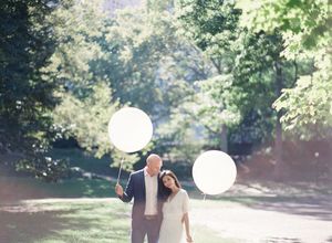 Couple standing close together holding large white balloons in a sunlit park pathway.