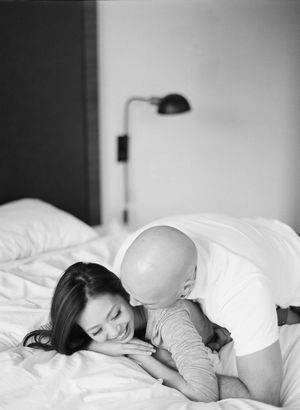A black and white photo of a man nuzzling a laughing woman lying on a white bed.