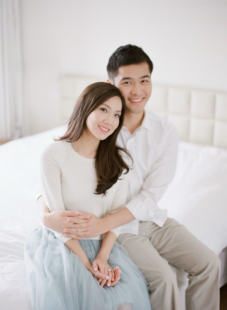 Smiling Asian couple sitting together on a white bed in a bright, airy room.