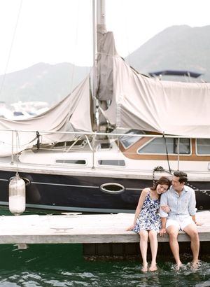 Romantic couple sitting on a dock with feet in water beside a sailboat.