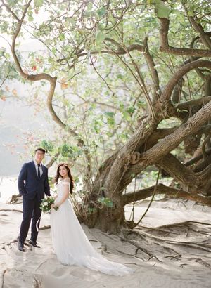 Wedding couple posing under a large twisted tree on a sandy beach with lush foliage.