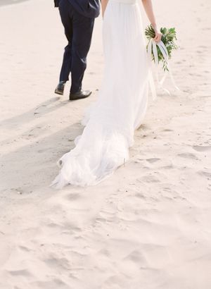 Close-up of bride walking in flowing white gown on sandy beach holding a bouquet.