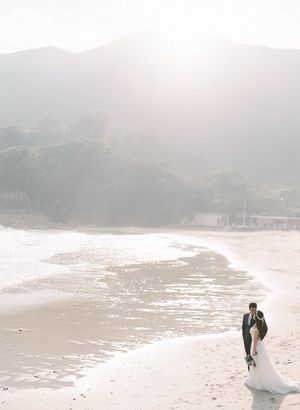 Bride and groom sharing a romantic moment on a misty sunlit beach with mountain backdrop.