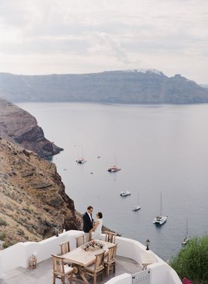Couple sharing romantic moment on cliffside terrace overlooking Santorini caldera with sailboats and dramatic sea views.