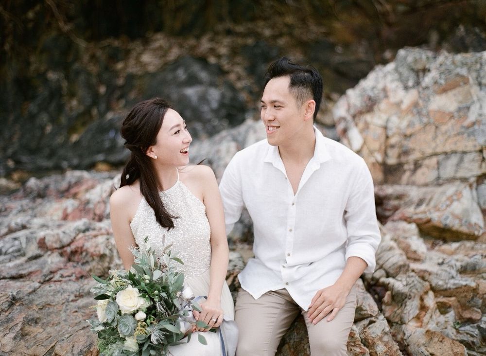 Happy couple sitting on rocky coastal landscape, smiling at each other with bridal bouquet of white flowers.