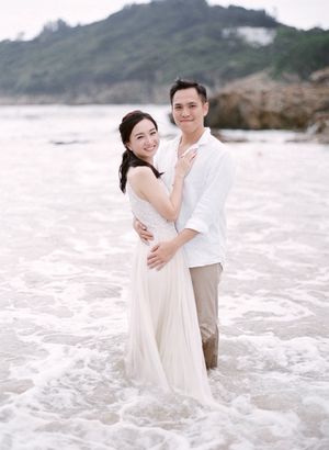 Joyful couple embracing in ocean waves, bride wearing flowing white dress during romantic beach photo session.