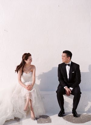 Elegant bride and groom sitting against white wall in Santorini, sharing a joyful candid moment together.