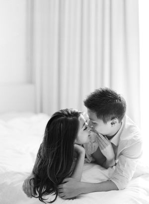 Tender black and white portrait of couple sharing a gentle kiss while lying together on white bedding.