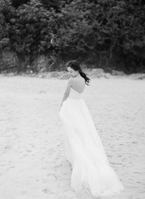 Black and white photo of a bride in a flowing gown walking on a sandy beach.