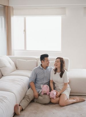 Happy couple sitting on floor against white sofa holding pink piggy banks indoors.