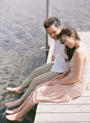 Couple sitting on a wooden dock with feet over water, smiling and embracing outdoors.