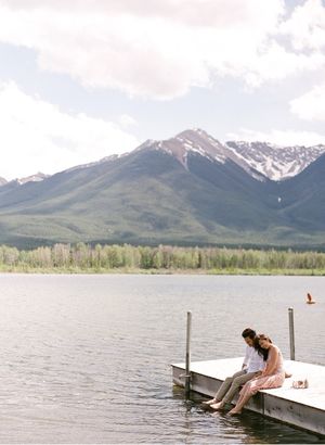 Couple sitting on a dock by a mountain lake with snow-capped peaks in background.