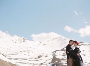 Couple embracing in front of dramatic snow-covered mountains under a bright blue sky.