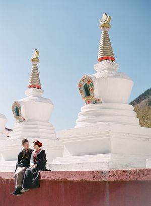 Couple sitting together in front of white Buddhist stupas with golden spires in traditional Tibetan attire.