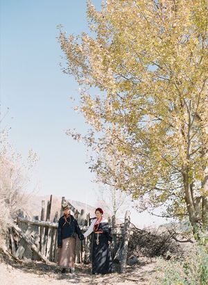 Couple in traditional Tibetan clothing walking near a rustic wooden fence beneath golden autumn trees.