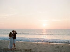 Couple dancing romantically on a beach at sunset with pastel pink sky and calm ocean waves.