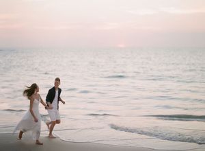 Joyful bride and groom running hand in hand along the shoreline at a pastel sunset beach.