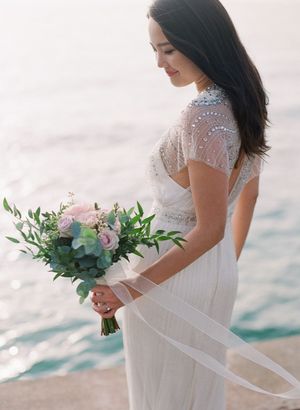 Bride in beaded white gown holding pink rose bouquet by the ocean at sunset.