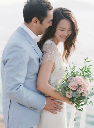 Groom in light blue suit embracing smiling bride holding pink roses bouquet near sparkling water.