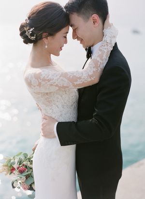 Bride in lace long-sleeve gown and groom in tuxedo embracing near shimmering water.