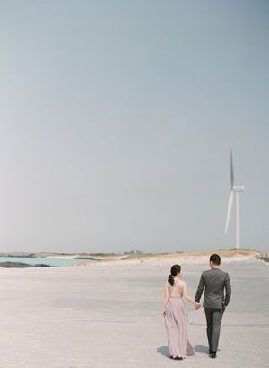 Couple holding hands walking on white sand beach with wind turbine in background.