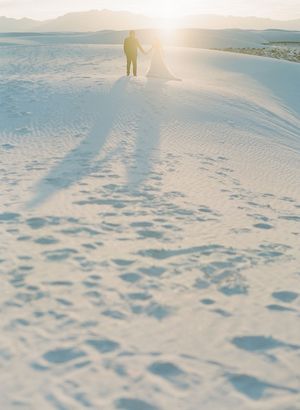 Couple silhouetted against golden sunset on white sand dunes, casting long shadows across the rippled desert landscape.