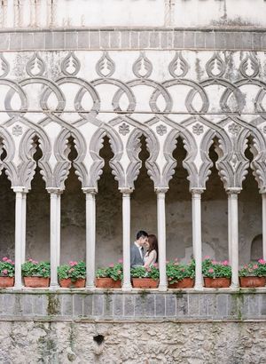 Couple shares romantic kiss framed by ornate Gothic arched colonnade with terracotta flower pots in historic Italian villa.