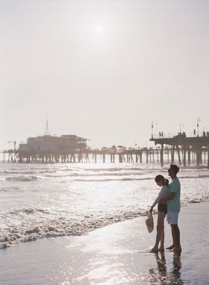 Couple stands at ocean's edge in silhouette with Santa Monica Pier glowing in bright hazy sunlight behind them.