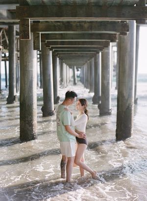 Couple embraces romantically under a pier surrounded by ocean waves and rows of wooden support columns.