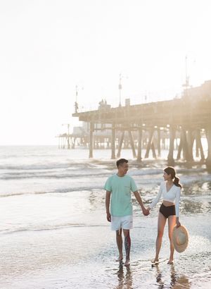 Couple holding hands walking along the beach near a pier during golden hour sunset.