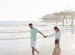 Young couple strolling hand in hand on a sunny beach with a pier in the background.