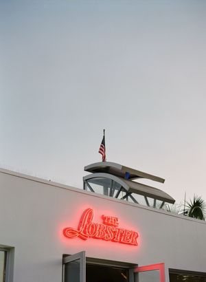 Neon red sign of The Lobster restaurant glows at dusk with American flag on rooftop against pale evening sky.