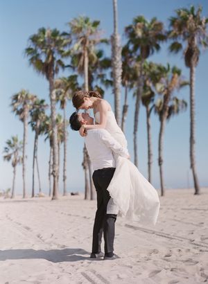 Groom lifts bride in white wedding dress for a kiss on sandy beach lined with tall palm trees under blue sky.